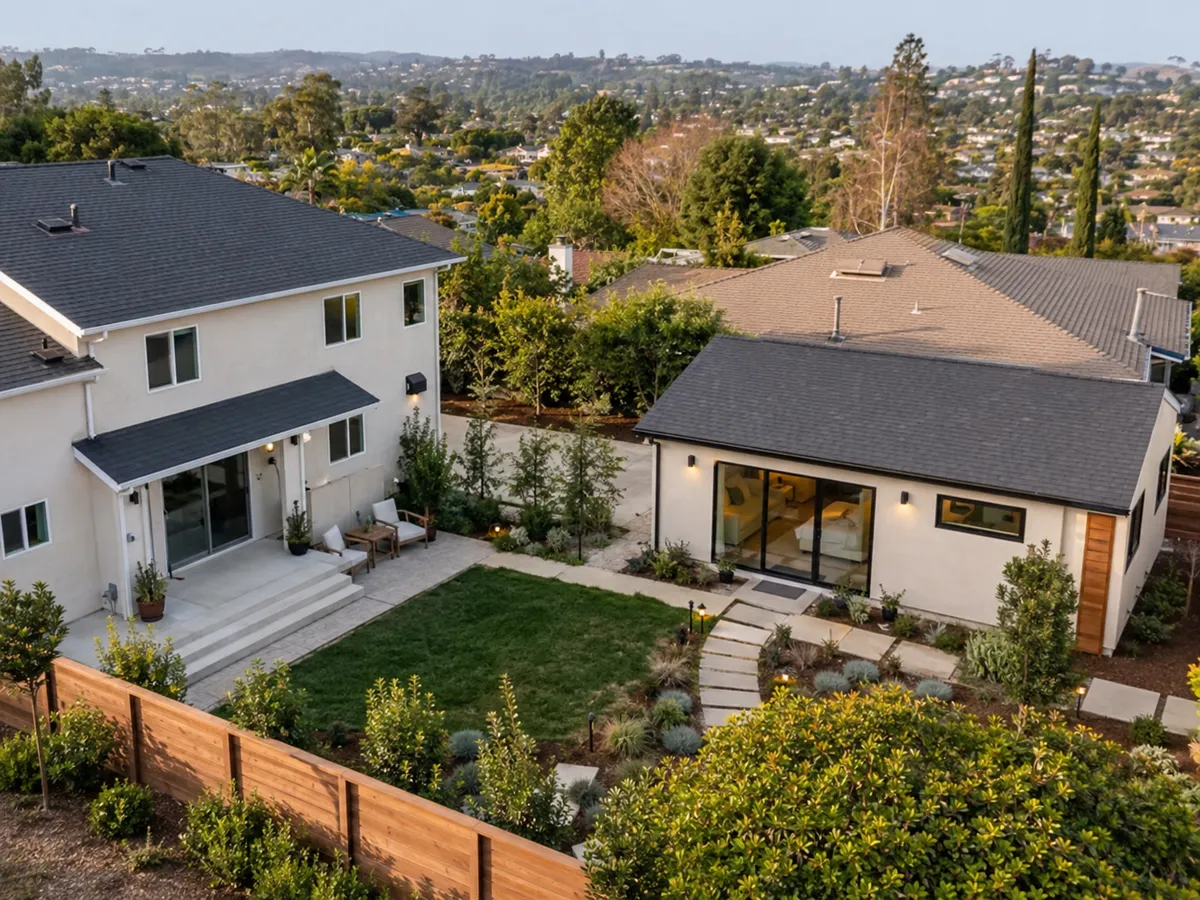 Aerial view at dusk of a completed San Diego County detached ADU next to the primary two-story house, showing the relative scale, shared yard, mature landscaping, and hillside suburban context